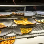 Thin-crust pizza slices on round metal trays in a lit display case, ready to be sold by the slice