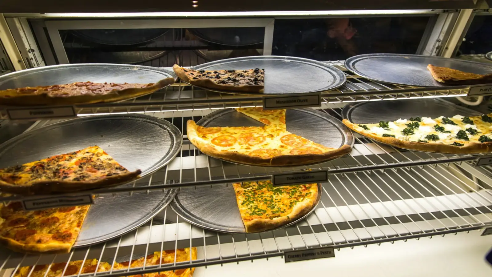 Thin-crust pizza slices on round metal trays in a lit display case, ready to be sold by the slice