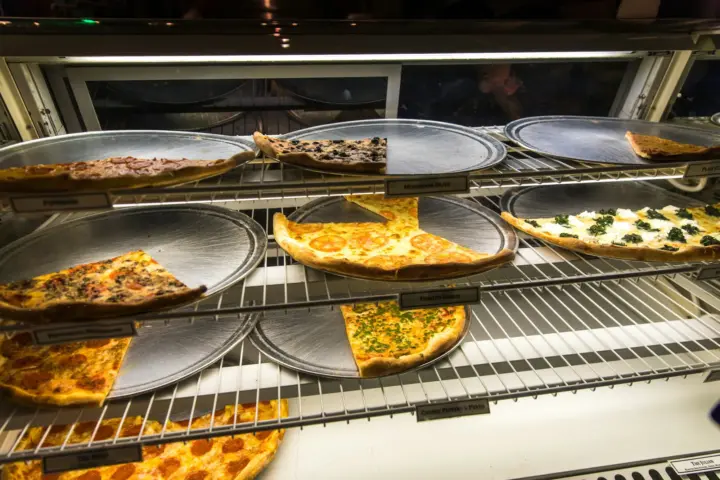 Thin-crust pizza slices on round metal trays in a lit display case, ready to be sold by the slice