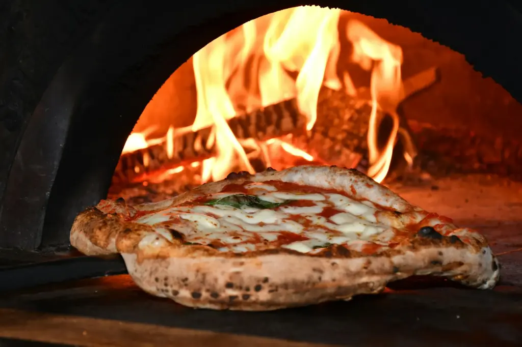 A margherita on a peel at the mouth of a wood-fired oven, logs burning brightly behind it