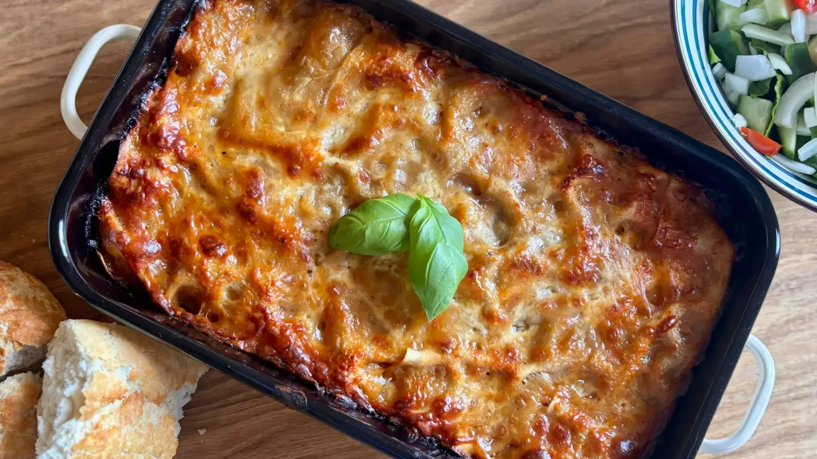 Golden, cheese-topped pasta bake in a dark rectangular baking dish with a basil leaf, crusty bread and salad