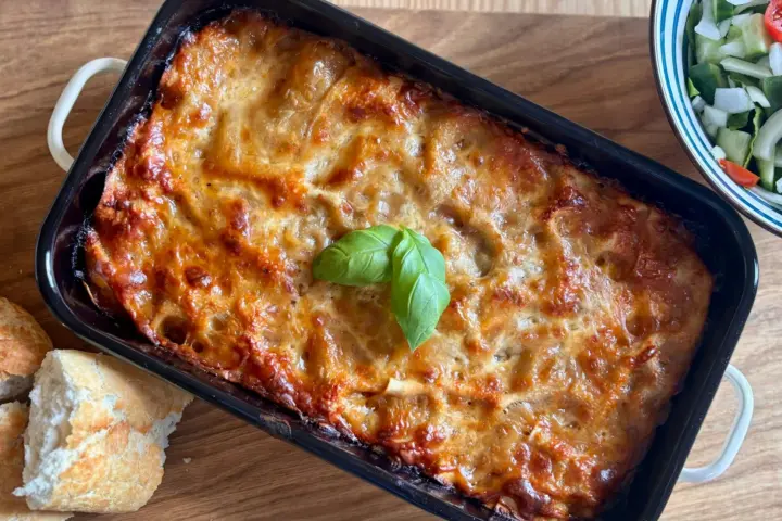 Golden, cheese-topped pasta bake in a dark rectangular baking dish with a basil leaf, crusty bread and salad