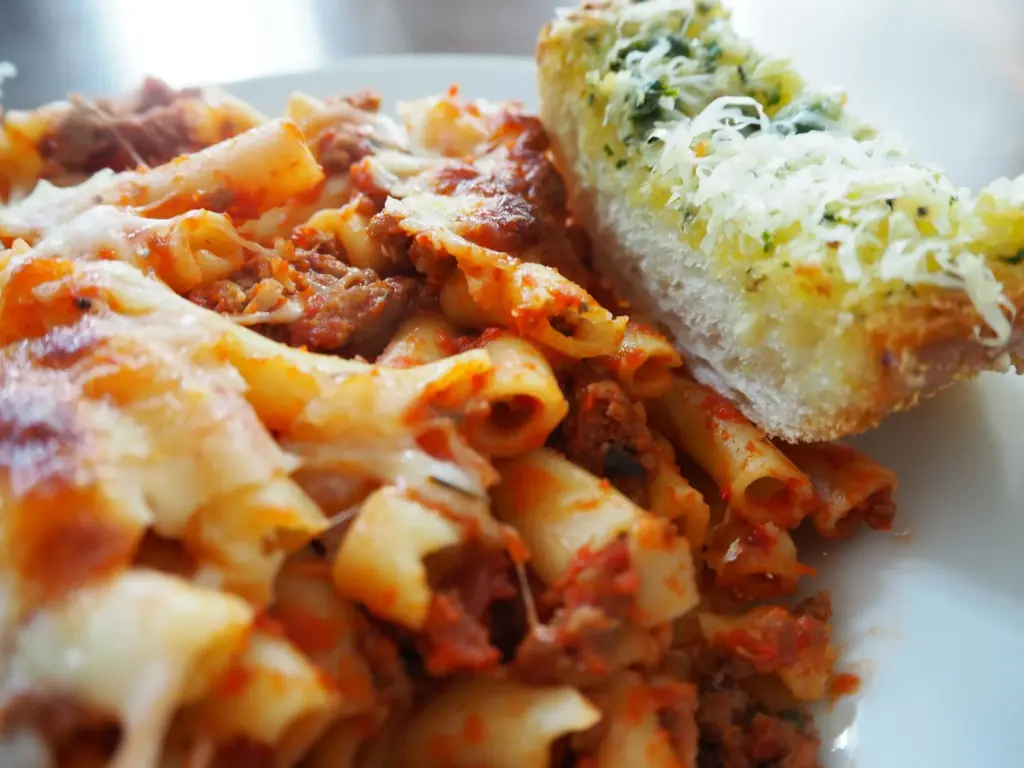 Plated baked ziti with meat sauce and browned, melted mozzarella next to a slice of cheesy garlic bread