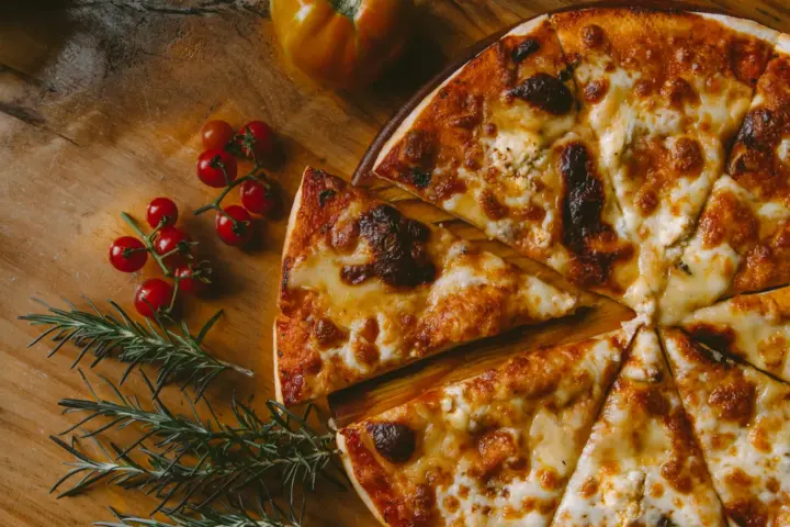 Sliced cheese pizza with browned, blistered mozzarella on a wooden board beside cherry tomatoes and rosemary