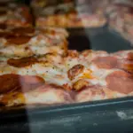 Square pan pizza with pepperoni and melted cheese, seen through the glass of a display case