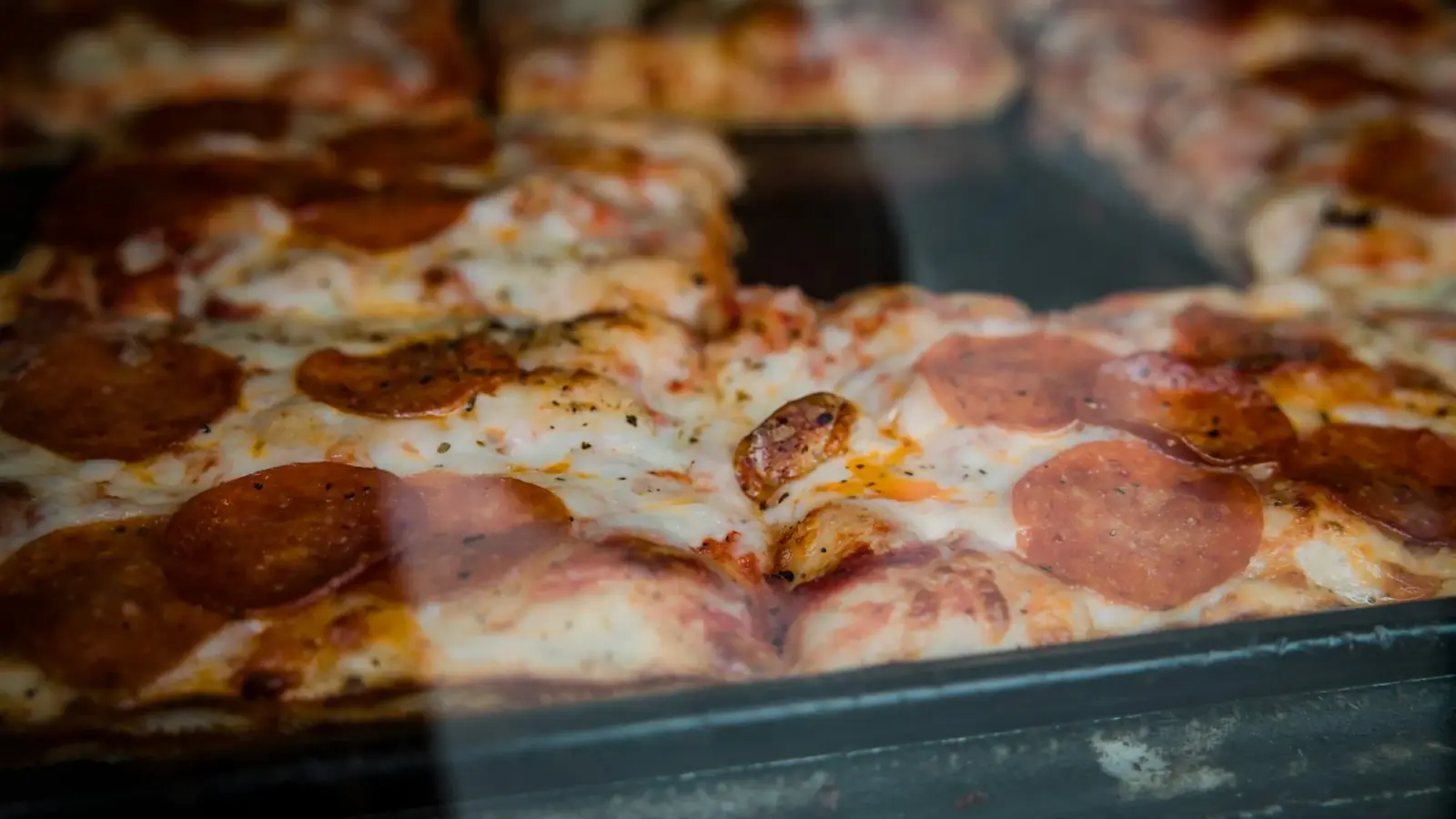 Square pan pizza with pepperoni and melted cheese, seen through the glass of a display case