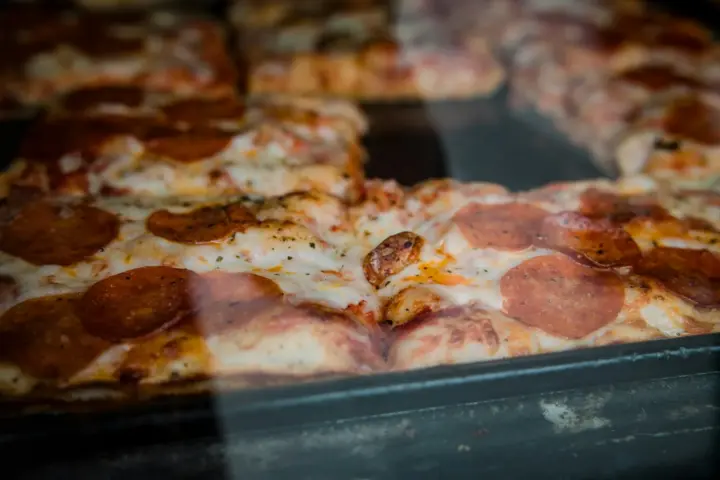 Square pan pizza with pepperoni and melted cheese, seen through the glass of a display case