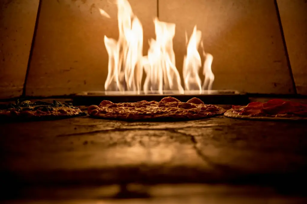 Thin pizzas baking directly on the stone floor of a gas-fired oven, with flames rising at the back