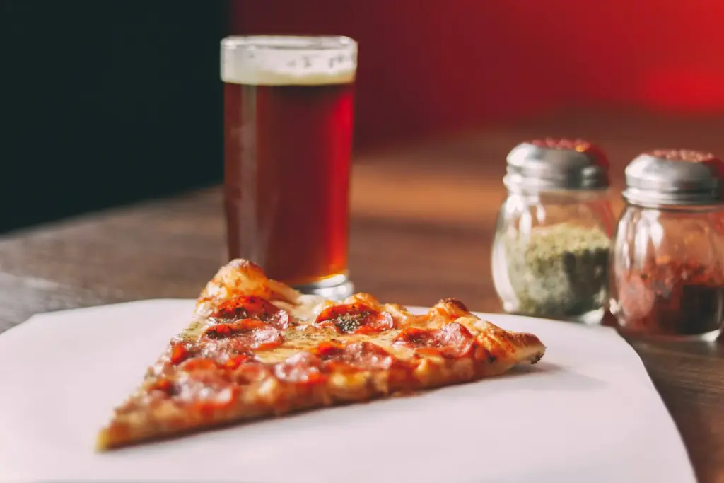 Pepperoni slice on white paper beside glass shakers of dried oregano and red pepper flakes and a glass of beer