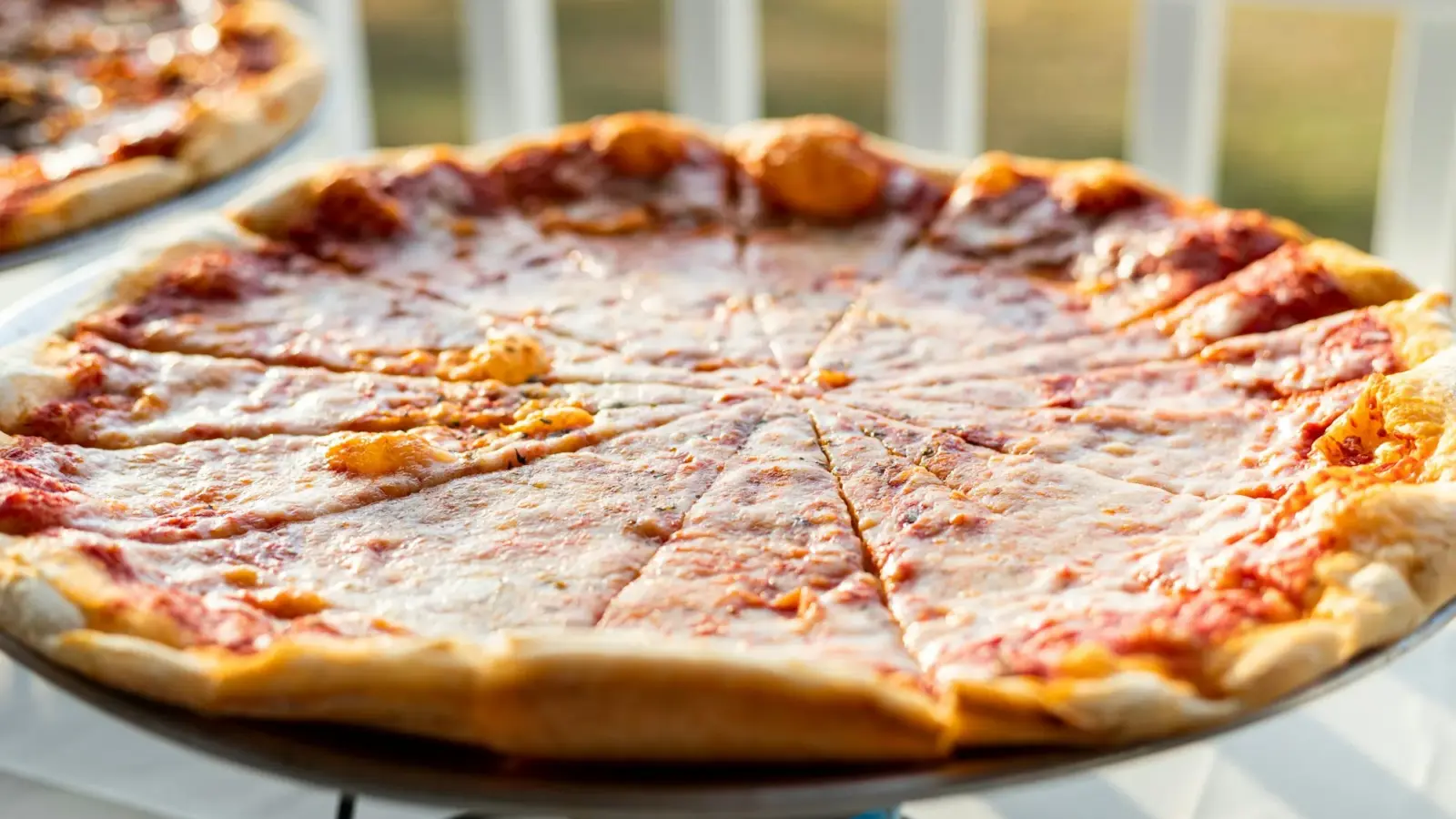 Large thin-crust cheese pizza cut into wedges on a raised metal pizza stand in warm afternoon light