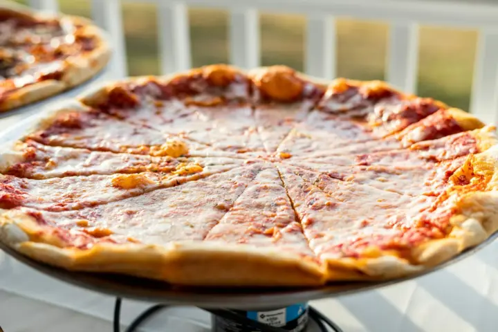 Large thin-crust cheese pizza cut into wedges on a raised metal pizza stand in warm afternoon light