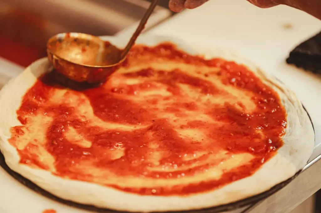 Ladle spreading a thin layer of tomato sauce over stretched pizza dough, leaving a bare border at the rim