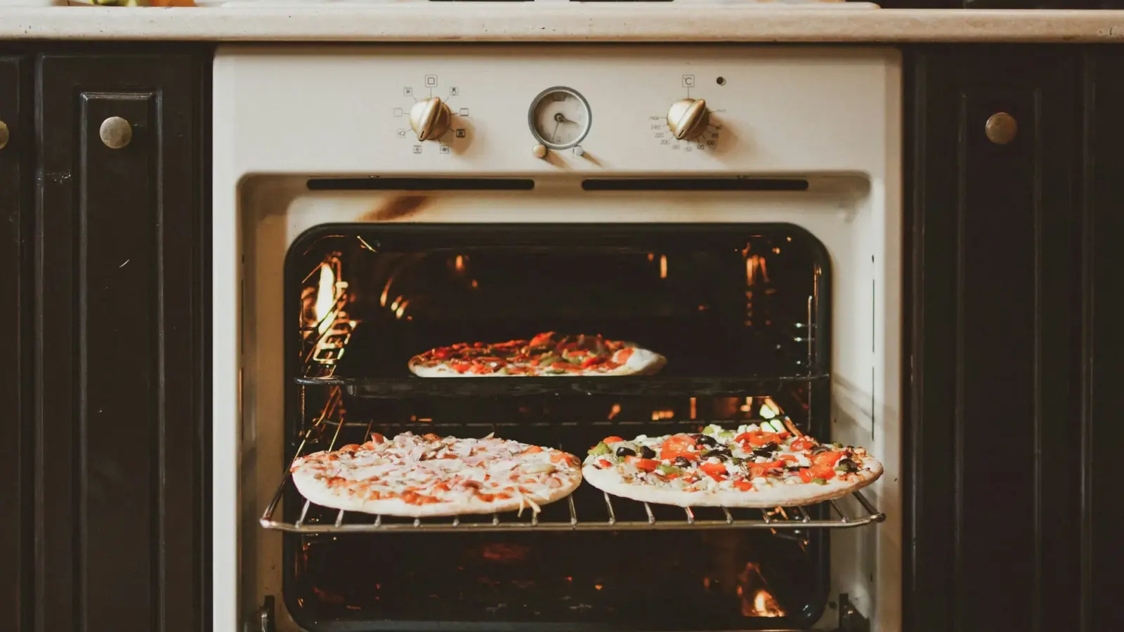 Three homemade pizzas baking on two racks of an open home oven, the heating element glowing at the top