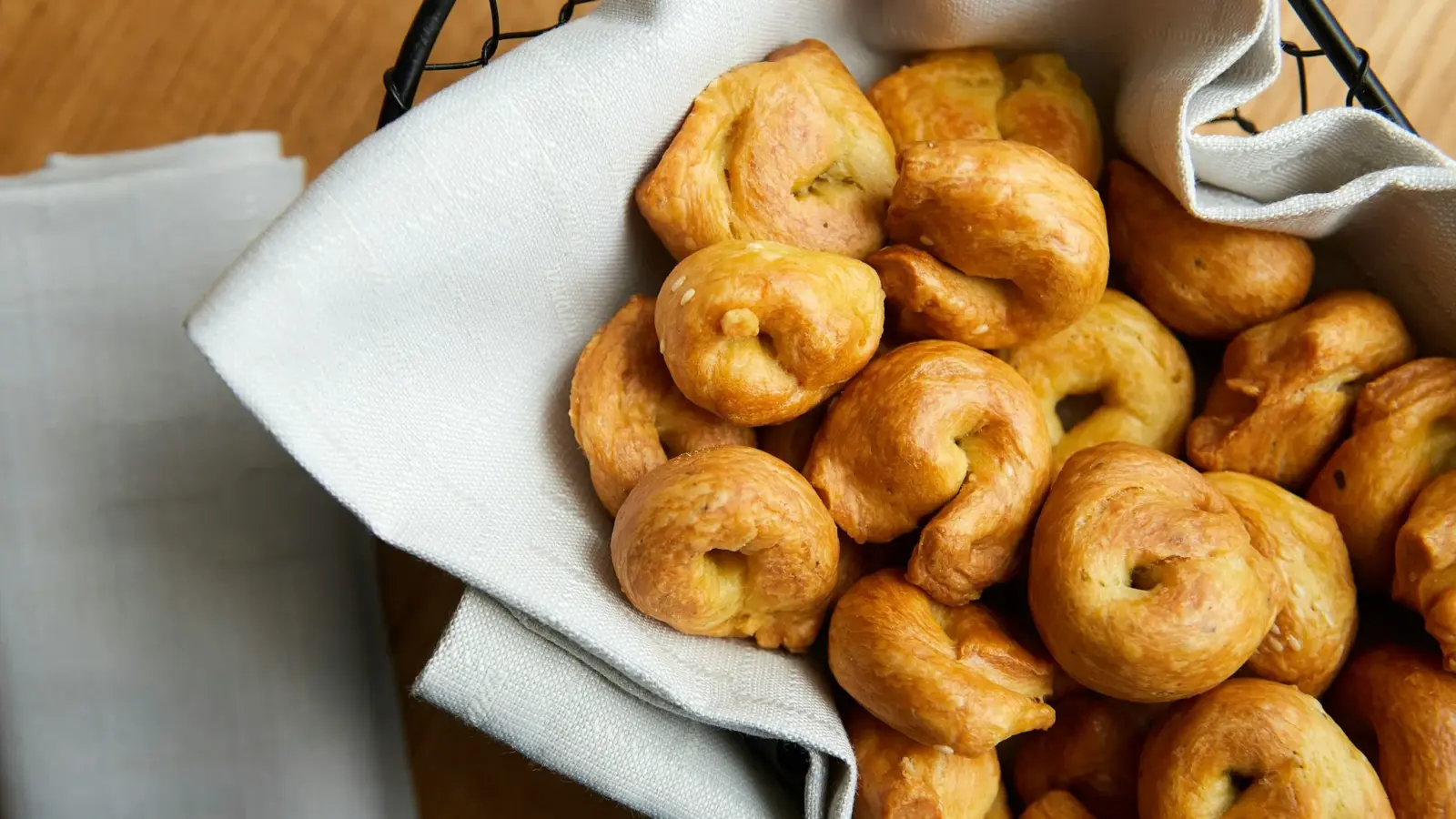 Small glossy knotted bread rolls piled in a wire basket lined with a linen napkin on a wooden table