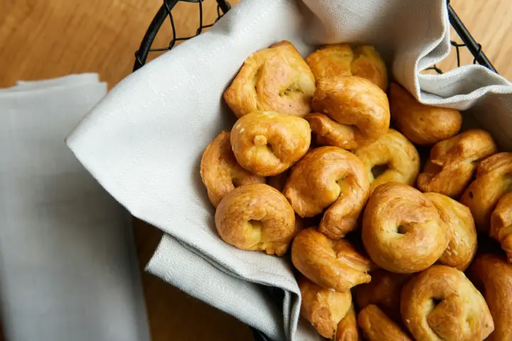 Small glossy knotted bread rolls piled in a wire basket lined with a linen napkin on a wooden table