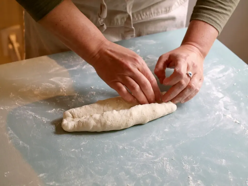 Hands shaping a piece of dough into a log on a lightly floured countertop