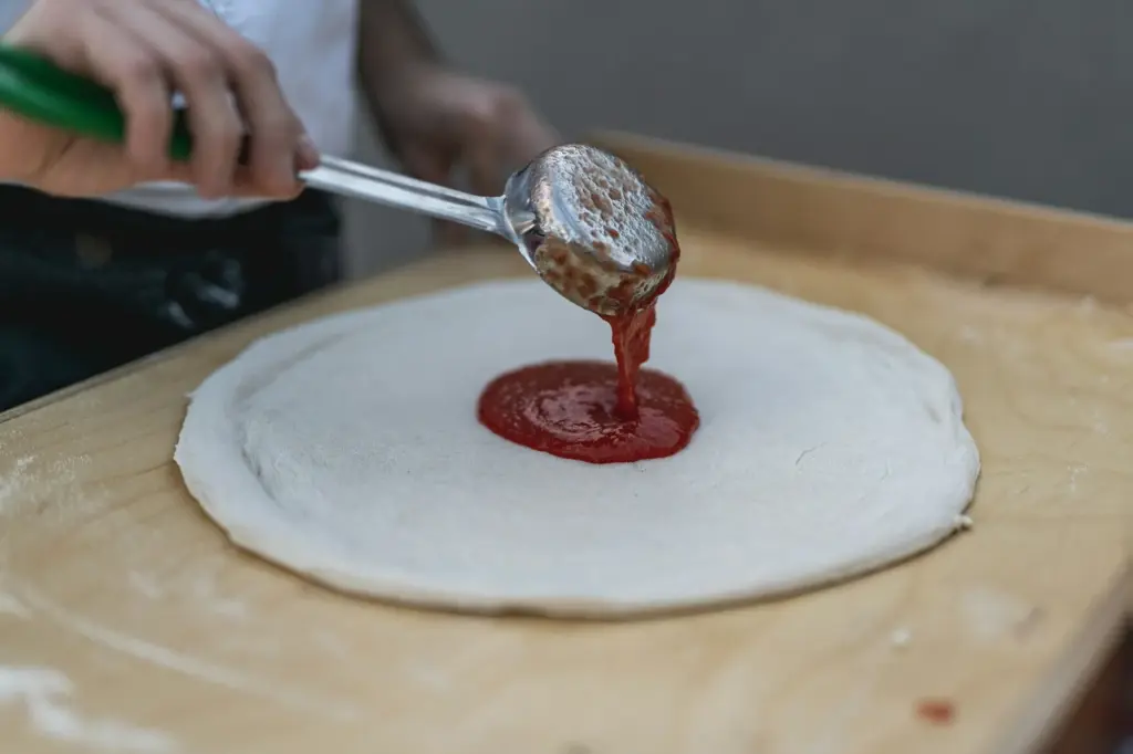 Tomato sauce poured from a ladle onto the center of stretched pizza dough on a floured wooden counter