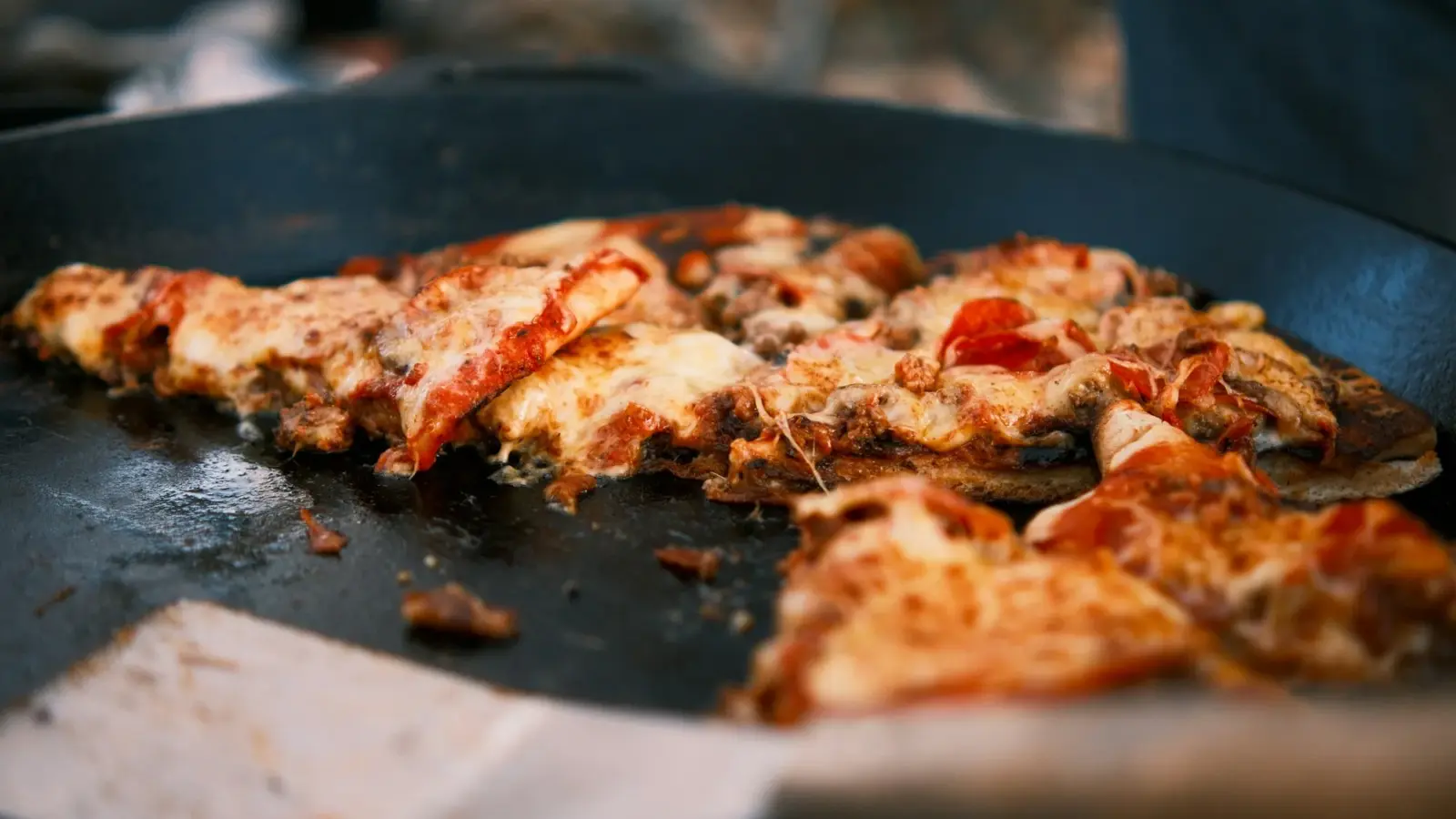 Thin-crust pizza pieces with bubbling cheese warming in a wide black pan, a metal spatula sliding in at the front