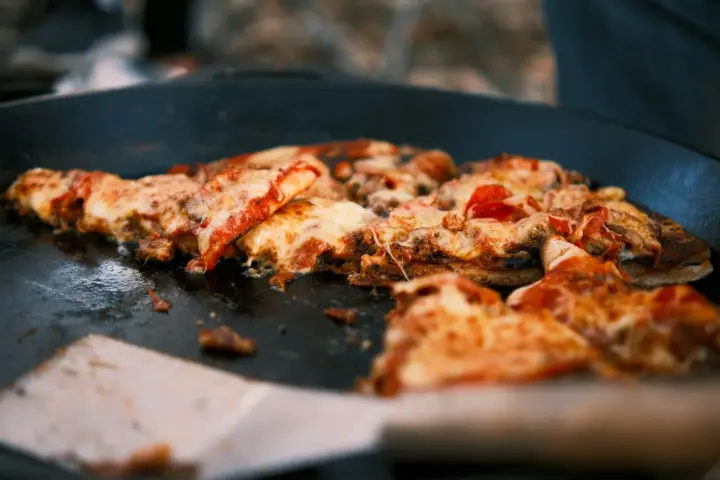Thin-crust pizza pieces with bubbling cheese warming in a wide black pan, a metal spatula sliding in at the front