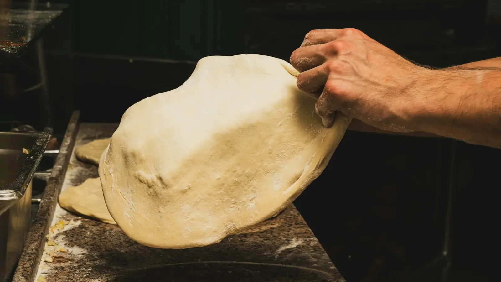A hand lifts a large, thin round of stretched pizza dough off a floured stone counter in a dim kitchen
