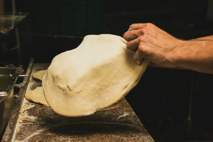 A hand lifts a large, thin round of stretched pizza dough off a floured stone counter in a dim kitchen