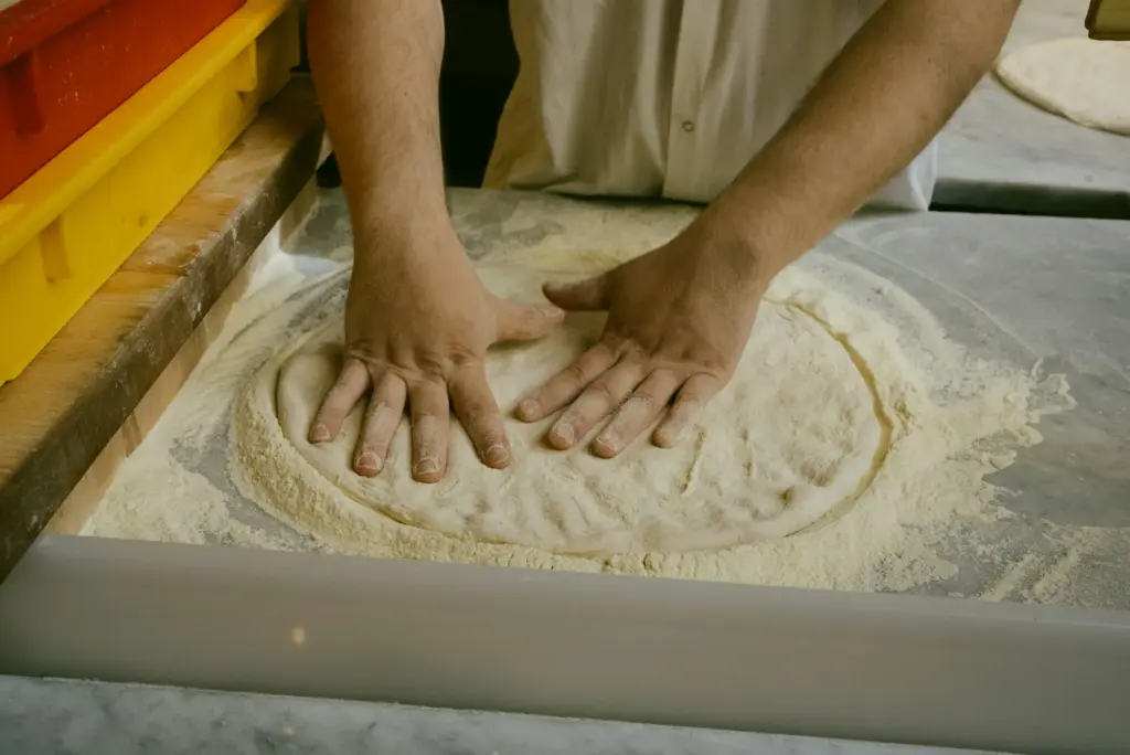 Fingertips press a round of dough flat on a heavily floured counter, leaving a thicker rim around the edge
