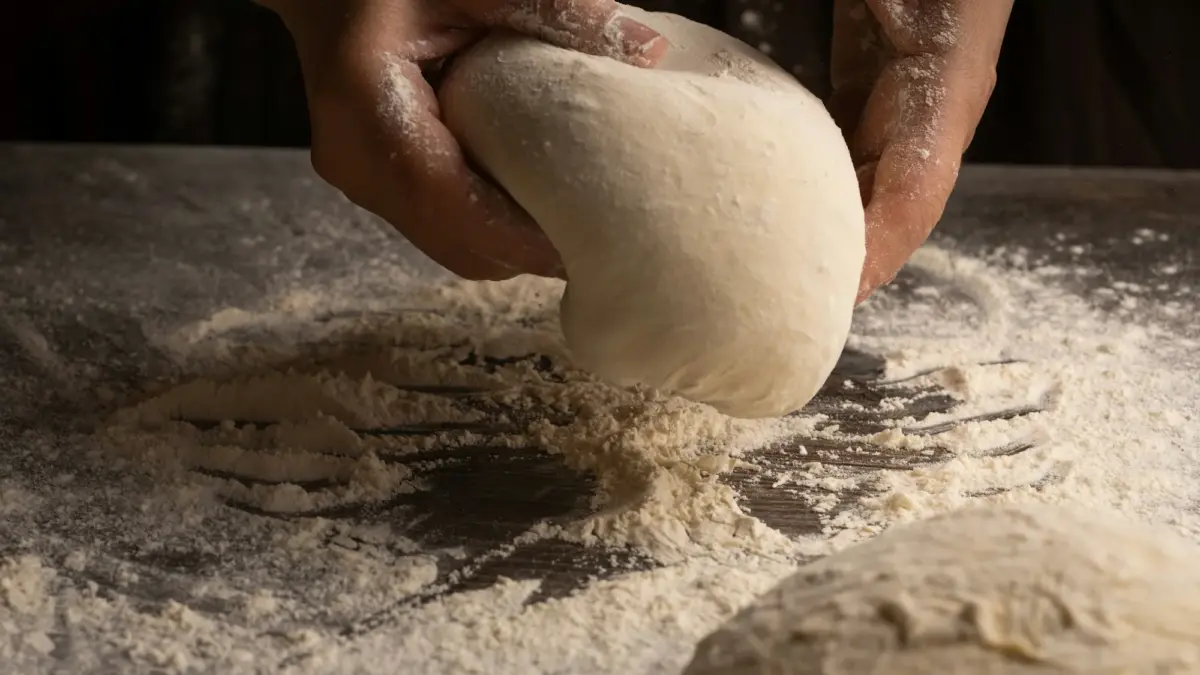 Floured hands lifting a smooth ball of pizza dough off a flour-dusted counter, a second dough ball in front