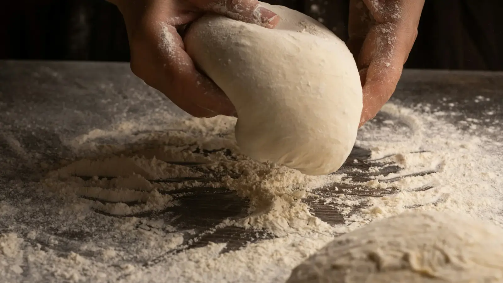 Floured hands lifting a smooth ball of pizza dough off a flour-dusted counter, a second dough ball in front