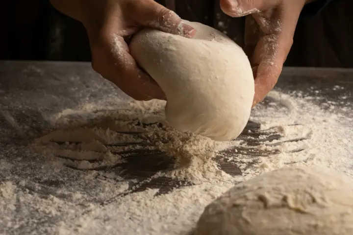 Floured hands lifting a smooth ball of pizza dough off a flour-dusted counter, a second dough ball in front