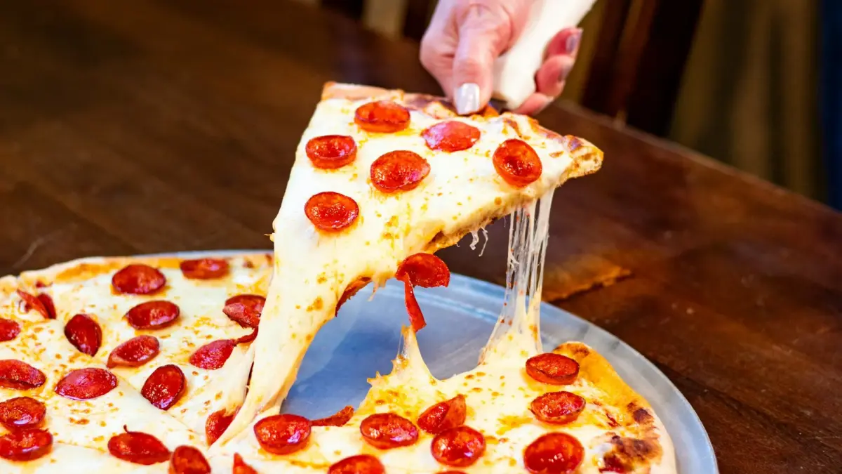 Hand lifting a slice from a thin-crust pepperoni pizza on a metal tray, melted cheese stretching