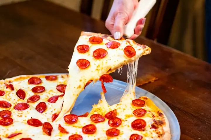 Hand lifting a slice from a thin-crust pepperoni pizza on a metal tray, melted cheese stretching