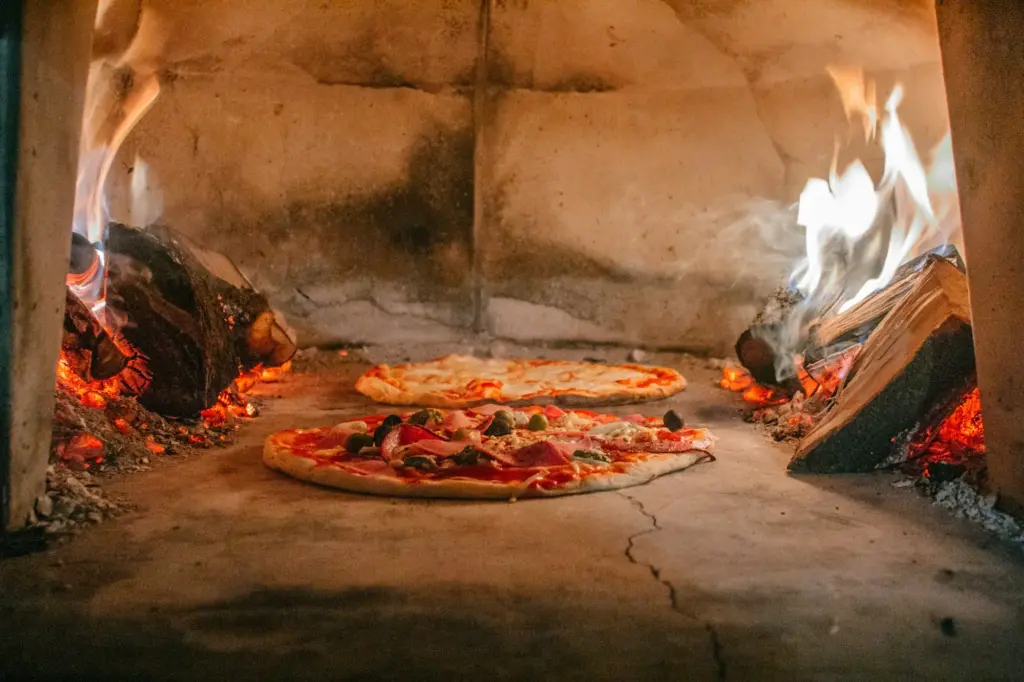 Two pizzas baking on the floor of a large stone oven between glowing fires
