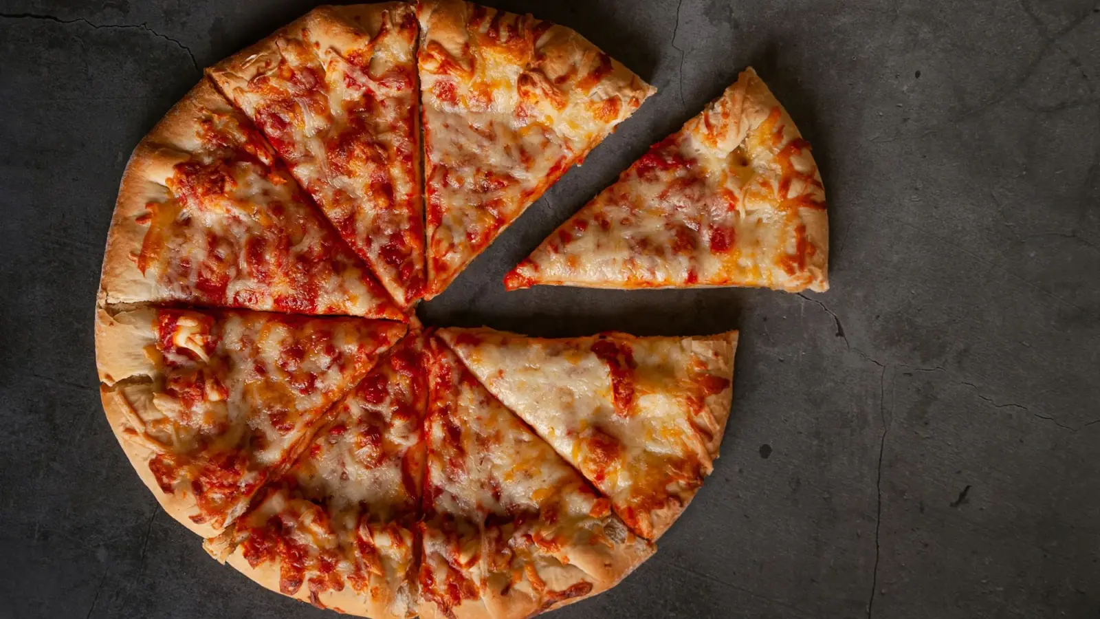 A thin-crust cheese pizza cut into eight slices on a dark slate surface, with one slice pulled away