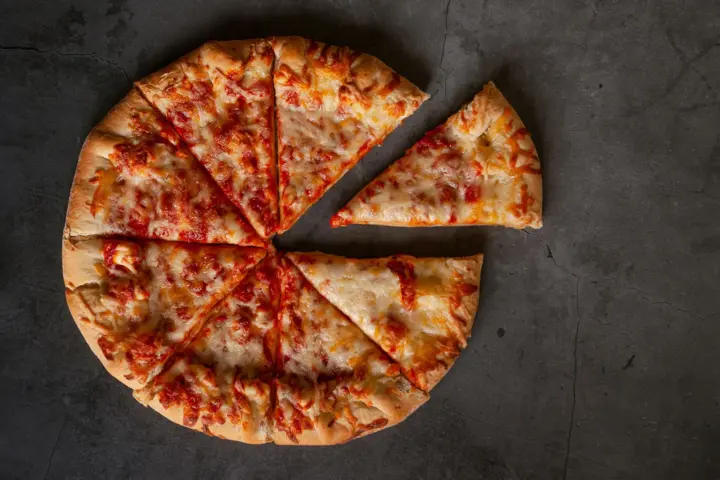 A thin-crust cheese pizza cut into eight slices on a dark slate surface, with one slice pulled away