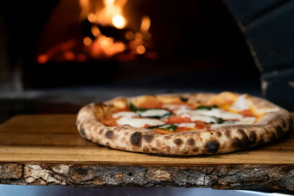 Low-angle view of a margherita's tall, puffy rim with small dark blisters, on a board before a wood-fired oven