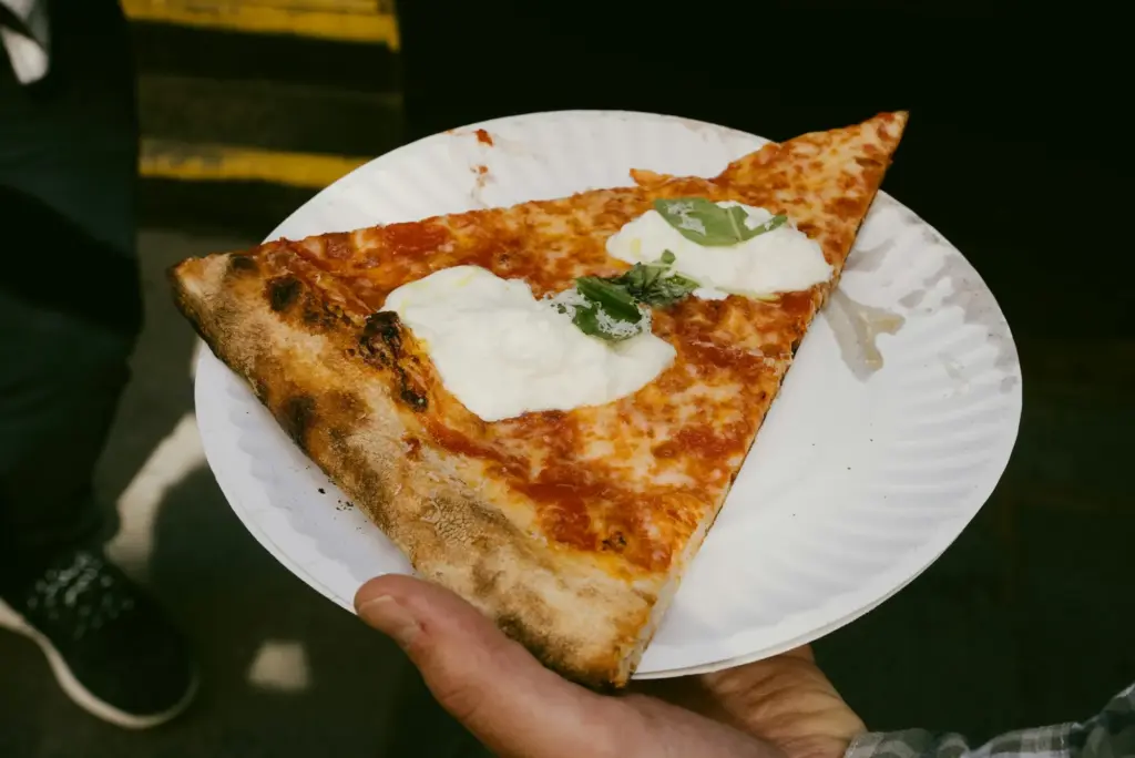 A hand holding a large, thin-crust pizza slice topped with fresh mozzarella on a white paper plate