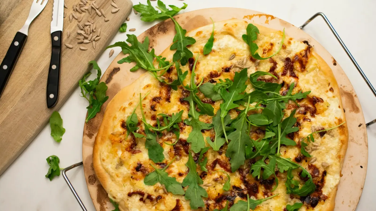 White pizza topped with arugula on a round, darkly stained pizza stone in a wire carrier on a marble counter