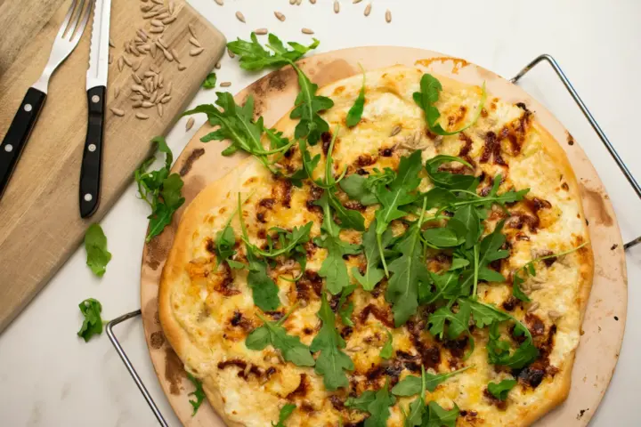 White pizza topped with arugula on a round, darkly stained pizza stone in a wire carrier on a marble counter