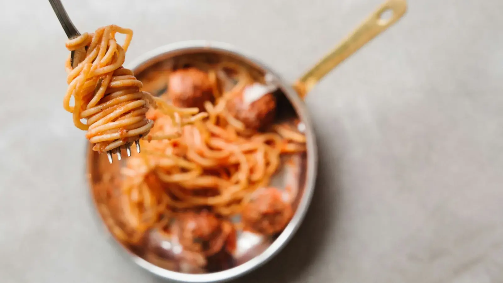 Fork twirling sauce-coated spaghetti above a small copper-handled pan of spaghetti and meatballs in tomato sauce
