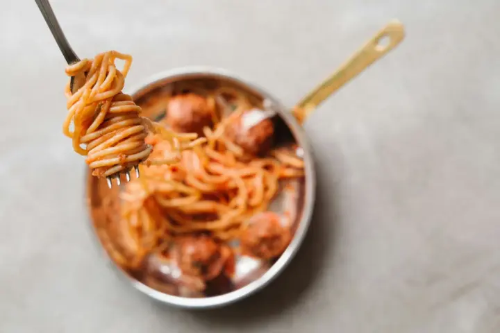 Fork twirling sauce-coated spaghetti above a small copper-handled pan of spaghetti and meatballs in tomato sauce