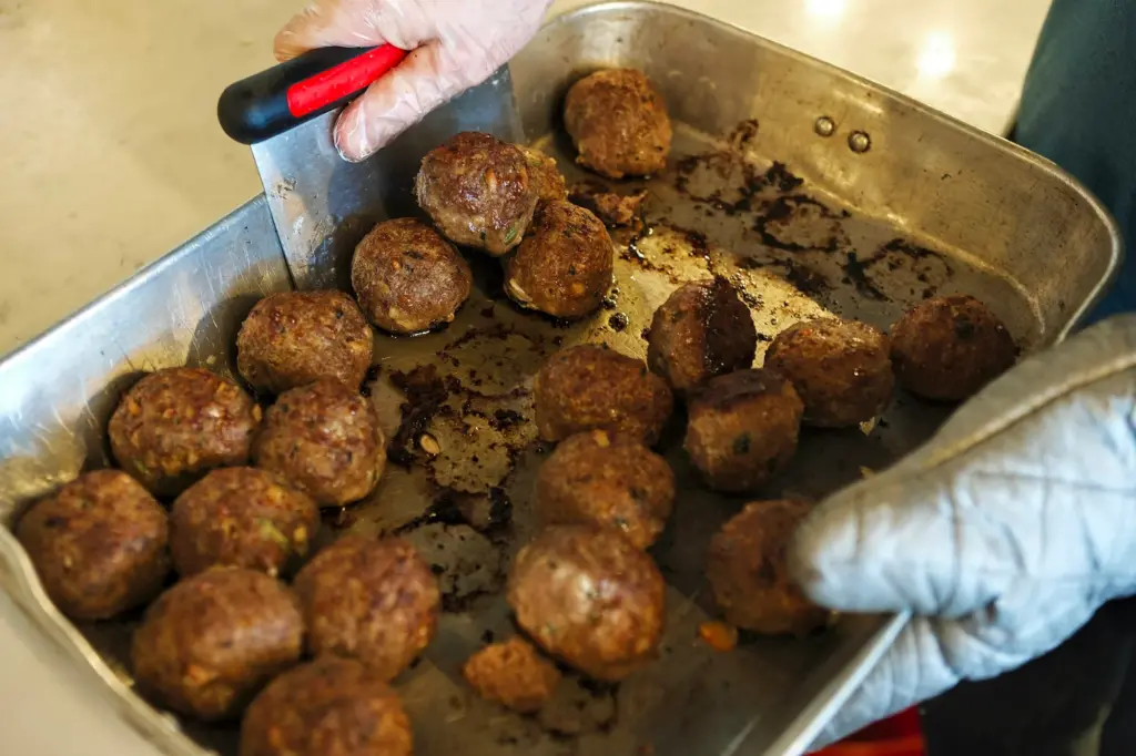 Gloved hands holding a metal roasting pan of browned meatballs with dark crusts and browned drippings